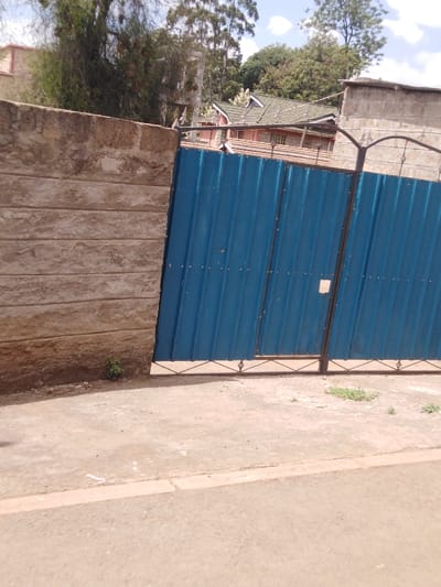 The image depicts a property boundary in Sigona ward, Kenya. In the foreground, an unpaved light brown ground surface transitions to a darker paved road separated by a low curb. A closed, two-panel gate made of blue corrugated metal with a black steel frame occupies the central and right portions of the frame. The gate features exposed rivet fasteners and a small light-colored square patch near the center-right. Barbed wire or razor wire is affixed along the top of the gate's frame. To the left of the gate, a section of a light-colored concrete block wall with horizontal tooling is visible. A small green plant grows at the base of this wall. In the mid-ground, behind the gate and wall, a residential building with a red-tiled roof and light-colored walls is partially visible, surrounded by dense green foliage and tall trees. A concrete structure is also visible behind the right portion of the gate. The sky above is light blue with scattered white clouds. No individuals or discernible actions are present in the scene.