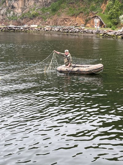 A person in camouflage clothing and a white cap is in an inflatable boat on the water, holding a fishing net with fish. The boat is camouflaged and has oars. In the background, there is a rocky cliff with green vegetation. Along the shoreline, there is a stone wall, and a small wooden building with a red brick chimney. The water is choppy, with reflections of the sky and surrounding environment. The location is the Bay of Sovetskaya Gavan', Russia.
