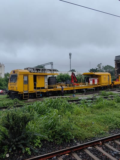 In Bhopal, India, on a cloudy day, two yellow railway maintenance trains are parked side-by-side on tracks overgrown with lush green vegetation. The train in the foreground is more extensively depicted, featuring a cabin with the text "8W आर बी एम टी MFG-2025" and "INDIAN RAILWAYS" visible on its side. The train is equipped with machinery, including a red crane at the rear of the first section and a red and white generator on the second section. A person stands near the front of the second section, looking towards the train. In the background, a cell tower stands against the overcast sky, and a building is partially visible. The scene suggests a maintenance yard or a work site for the Indian Railways.