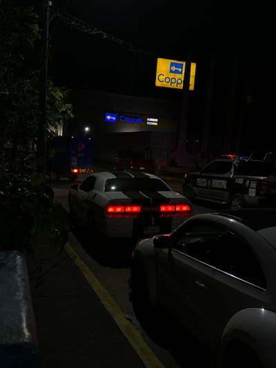 At night, a Coppel store sign shines brightly with its key logo. Behind it, multiple vehicles are parked along the curb. A white car is prominent, its taillights glowing, parked near a curb marked with a yellow line. A police vehicle is also present. Lush greenery surrounds the area, with a Pepsi sign in the background, partly obscured by the foliage. The dark setting contrasts with the lit signs and car lights.