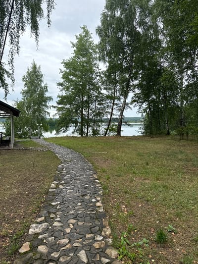 The image depicts a stone pathway leading towards a serene lake surrounded by lush green trees. The pathway, made of irregular grey stones, curves gently to the right, disappearing into the distance. To the left of the pathway, a wooden structure resembling a gazebo or pavilion is visible, with seating arrangements inside. Beyond the pathway and to the left, the lake stretches out, reflecting the overcast sky. The trees, predominantly birch with their characteristic white bark, line the shore of the lake and fill the background, creating a dense, natural setting. The sky is cloudy, suggesting a cool or temperate day. The overall scene evokes a peaceful, natural environment, likely a recreational area. This location is identified as being in the city of Leninskii, Russia.