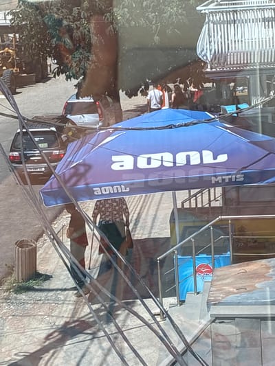 The image shows an outdoor scene in Khashuri, Georgia, viewed through a window with reflections. In the foreground, a blue canopy with white text, "MTIS" is visible. Two people are partially obscured by the canopy, one wearing a patterned shirt and the other in a red and white striped top. In the mid-ground, a black car with a "TAXI" sign on its rear window is parked. Further back, a silver car is visible. The environment includes trees, power lines, and a construction vehicle in the distance. The ground appears to be paved with some debris and vegetation. A trash can is on the left side of the image.