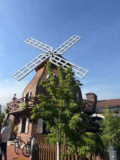 The image shows a picturesque windmill in the city of Seltso, Russia, under a clear blue sky. The windmill is constructed of brick and wood, with a shingled roof. Its large, white, lattice-like sails are angled against the sky. A wooden balcony with white railings and floral decorations adorns the side of the windmill. In the foreground, a person in a white shirt is walking past a brick building. Two bicycles are parked by the building, which has arched windows with white shutters and a wooden fence in front of it. Lush green trees and foliage surround the windmill and building, adding to the serene and picturesque atmosphere.
