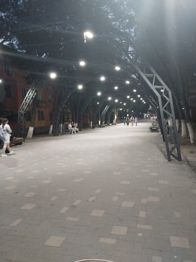 This image captures an outdoor scene at dusk, likely in the city of Ötegen batyr, Kazakhstan, focusing on a pedestrian walkway lined with trees and an overhead structure adorned with lights. The walkway is paved with grey tiles, some of which are lighter in color, creating a patterned effect. Along the sides of the walkway, there are benches and trees. An arched metal structure with a geometric design spans overhead, from which numerous round lights are suspended, illuminating the pathway. In the background, a building with orange walls and dark windows is visible, suggesting an urban or semi-urban setting.

Several individuals are present in the scene. On the left side of the walkway, a person wearing a light-colored dress and white shoes is walking away from the camera. Further down the walkway, on the left, two people are seated on a bench. Towards the middle ground, a group of three people, two adults and a child, are walking together. In the distance, more people can be seen strolling along the path, indicating a public space that is being utilized by residents. The overall atmosphere is calm and the lighting suggests the transition from day to night.