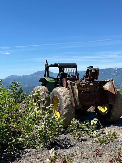 An abandoned, rusty, and weathered farm tractor sits on a gravelly, mountainous terrain in Crkvine, Montenegro. The tractor, predominantly green and brown with yellow accents on its wheels, is positioned at an angle, facing right. Its large, mud-caked tires are prominent. Vegetation, consisting of leafy green plants and shrubs, grows around the tractor and in the foreground, some of which appears to be encroaching on the machinery. In the background, a vast mountain range stretches across the horizon under a clear blue sky with a few wispy clouds and contrails. The overall scene suggests a remote, possibly disused, location within a natural, mountainous landscape.