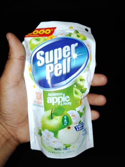 A person's hand is holding a pouch of Super Pell floor cleaner, possibly in Sendangrejo, Indonesia. The pouch is white with blue, green, and yellow accents. The brand name "Super Pell" is prominently displayed in large blue letters with a white outline. The scent is "Japanese apple & Peony." There is an image of green apples and white flowers on the pouch. Text on the pouch includes "10X LEBIH SEGAR" (10X FRESHER), "Quick Clean & Dry," and "Netto 100 g." The background is black.