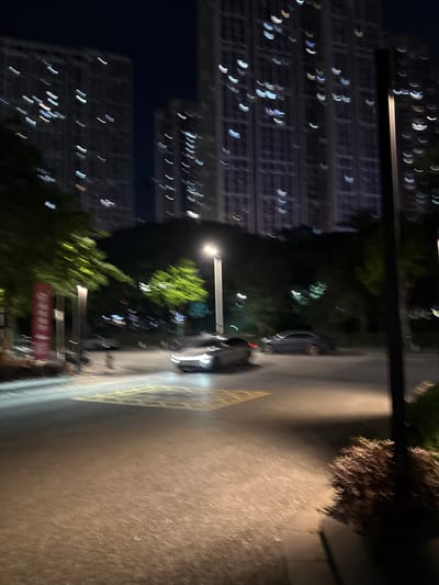 This image captures a blurry night scene in Haizhu District, China, focusing on an intersection with a paved road and sidewalk. Towering residential buildings, illuminated by numerous windows, dominate the background. Green trees line the area, their leaves blurred into streaks of light. A street lamp stands tall on the right side of the frame, casting a warm glow. Two cars are visible on the road, their forms indistinct due to motion blur. One car, a light-colored sedan with its headlights on, appears to be driving through a yellow-marked intersection. Another darker sedan is parked or moving slowly on the left side of the road. A red sign, also blurred, is positioned on the left side of the sidewalk. The overall impression is one of movement and the vibrancy of urban life at night, with the motion blur contributing to a dynamic and almost abstract aesthetic.