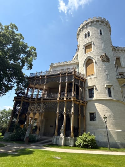 The image depicts the Hluboká Castle, a prominent landmark in Hluboká nad Vltavou, Czechia. The castle features a distinctive white stone structure with a circular tower, characteristic Gothic and Neo-Gothic architectural elements, and an ornate, multi-level balcony with intricate metalwork. The castle is situated on a manicured green lawn, with a pathway in the foreground and a large tree on the left side of the frame. The sky above is clear and blue with a few scattered clouds. A vintage-style lamppost stands to the right of the castle, casting a shadow on the building and the lawn. The scene is bathed in sunlight, indicating a daytime setting. There are no people visible in the image.
