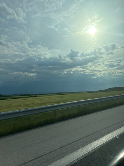 The image depicts a view of a sky with scattered clouds and a bright sun, alongside a stretch of green field, a metal guardrail, and a paved road. The road has a white painted line. The image appears to be taken from a moving vehicle. There is no evidence to definitively place the location in Zetiovo, Bulgaria.