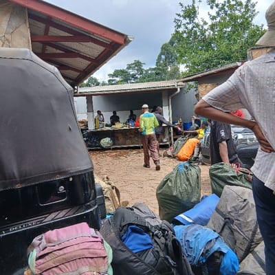 The image displays an outdoor gathering area in Mweka, Tanzania. In the immediate foreground, a dense accumulation of luggage, including backpacks, duffel bags, and large green utility sacks, is visible. To the left, the rear portion of a black three-wheeled vehicle, bearing the inscription "DELUXE," is partially obscured by luggage.

The midground features a reddish-brown paved surface, showing evidence of dampness. A man wearing a light green and yellow long-sleeved shirt, brown trousers, and a white cap walks away from the viewer, moving towards a single-story building in the background. This building, constructed with grey walls and a corrugated metal roof, hosts a long counter. Approximately five individuals, all appearing dark-skinned, are positioned behind and around this counter, engaged in various activities. One person wears a striped top, another a black shirt with a white graphic. Additional large bags, including an orange one and several green sacks, are situated on the ground in this area.

On the far right of the foreground, the torso and head of a man wearing a light-colored plaid shirt and a wide-brimmed hat are visible, with his hands on his hips. Architectural elements include an overhanging structure on the left with a corrugated metal roof supported by red-painted metal beams. The background is characterized by lush green trees and foliage beneath an overcast sky.