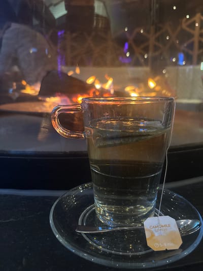 A clear glass mug, with a tea bag and spoon, sits atop a clear glass saucer. The tea bag is labeled "Camomile" and "5-8 min". The mug is filled with tea, with the tea bag inside. The background is blurry, with abstract shapes and bokeh lights. The mug sits on a dark, possibly black, surface.