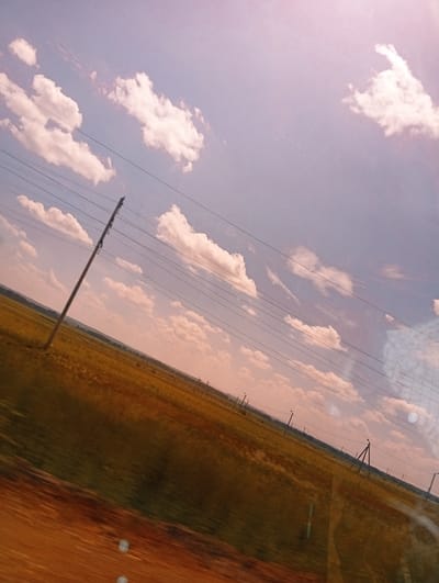 A tilted shot of a vast, sunlit field with tall grass under a pale blue sky filled with scattered, fluffy white clouds. Power lines stretch across the sky, supported by utility poles. One large, angled pole is prominent in the foreground. The horizon is low, showing a gentle slope of land. The overall lighting suggests it is daytime, possibly late afternoon or early morning due to the warm hues.