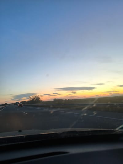 The image shows a view from inside a car driving on a highway at dusk. The sky transitions from blue at the top to orange and yellow near the horizon. There are some scattered clouds. The road is a multi-lane highway with white lines marking the lanes. Other vehicles, including a car and a truck, are visible in the distance. The landscape on either side of the highway appears to be fields and trees. The dashboard and windshield of the car are visible in the foreground. It is impossible to determine from this image if the location is in the city of Cherna Gora, Bulgaria.