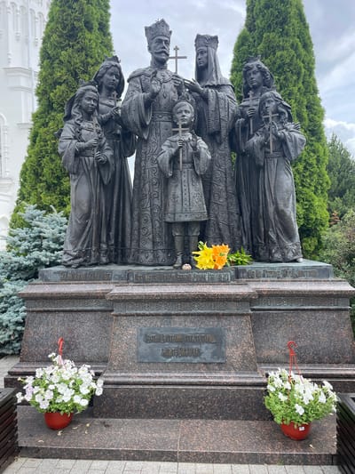 The image depicts a bronze sculpture of a family group standing on a granite pedestal in front of cypress trees and a white building, likely a church. The sculpture features a male and female figure at the center, presumably the parents, with four children arranged around them. All figures are adorned with crowns and hold crosses. The male figure wears a crown and a long, ornate robe. The female figure also wears a crown and a similar robe. The children, two boys and two girls, are dressed in historical attire and also hold crosses. A bouquet of orange and yellow flowers rests at the base of the sculpture. Two potted plants filled with white flowers are placed on the lower level of the pedestal. Inscriptions are visible on the pedestal, indicating names or historical references. The environment suggests an outdoor setting, likely a memorial site. The overall scene conveys a solemn and historical atmosphere, suggesting a monument dedicated to a significant family. The city of Diveyevo, Russia, is known for its religious sites, including the Diveyevo Monastery, which often features historical and religious monuments.