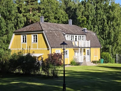 The image shows a bright yellow, two-story house with a dark gray, tiled roof, situated in Slaka, Sweden. The house features white-trimmed windows and a white-railed balcony on the upper level. There are two dark gray chimneys on the roof. The house is surrounded by lush green grass and mature trees, some of which are very tall and dense. In the foreground, a black lamppost stands on the lawn, and shadows from unseen trees are cast across the grass. Bushes and shrubs are visible along the side of the house. To the right of the house, a small wooden bench, a green trash bin, and a wire fence are present. The sky is clear and blue.