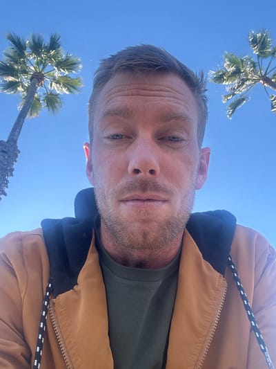 A man with short, light hair and light stubble takes a low-angle selfie against a clear blue sky. Palm trees are visible in the corners of the image. He is wearing a green t-shirt and a tan jacket with a dark hood. A black and white rope detail is visible on the jacket. The man's face is the primary focus, with his gaze directed toward the camera.