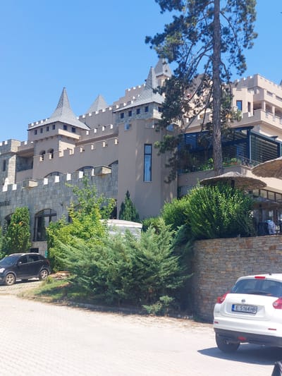 An exterior, daytime, eye-level shot features a resort-style building resembling a castle in Ognyanovo, Bulgaria. The building has a beige facade with stone accents, crenellated parapets, and several conical gray roofs. A large pine tree partially obscures the upper floors. In the foreground, a paved area is visible, with a dark SUV parked on the left and a white hatchback with a Bulgarian license plate "E 5164 HB" on the right. Lush green bushes and a stone wall separate the paved area from the building's lower level, where windows and an outdoor seating area with straw umbrellas are visible. The sky is clear and blue.