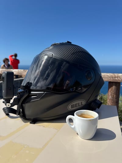 A matte black Bell motorcycle helmet, featuring a carbon fiber pattern on its crown and a dark tinted visor, rests on a light-colored, possibly tiled, outdoor table. An Insta360 action camera is mounted on the helmet's left side via a strap. A small white ceramic cup containing espresso is situated to the right of the helmet on the same table. The scene is set at a coastal viewpoint in Deià, Spain, overlooking the deep blue Mediterranean Sea under a clear blue sky. A rustic wooden railing runs horizontally across the mid-ground. Two blurred individuals are visible in the distant background behind the railing; one wears a red top and appears to be observing the sea.