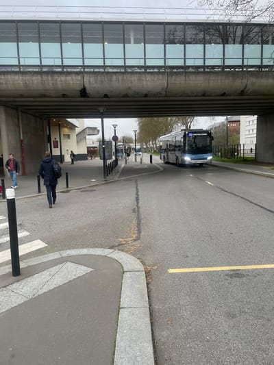 The scene depicts an urban transport area in Montigny-le-Bretonneux, France, specifically beneath an elevated pedestrian bridge near what appears to be a transit hub. The sky is overcast, providing diffuse lighting.
An elevated concrete structure with a glass-paneled pedestrian walkway spans the upper portion of the image, forming an underpass. To the left, a multi-story building facade with an entrance is visible under this structure.
The ground consists of a grey asphalt road that curves slightly to the right, extending into the mid-ground. A yellow line marking is present on the asphalt. To the left of the road, a grey paved pedestrian sidewalk is defined by granite curbs and black bollards. A marked pedestrian crosswalk with white stripes is visible in the foreground on the left.
A blue and white public transit bus, displaying "SNCF Pôle Paris Viroflay" on its destination sign and with illuminated headlights, is positioned on the right side of the road, moving towards the viewer. A second, similar bus is partially visible in the distant background on the right.
Three individuals are present. One male, wearing a dark blue jacket, dark trousers, and carrying a backpack, walks away from the viewer on the left sidewalk. Another male, wearing a red jacket and blue jeans, walks towards the viewer on the far left. A female subject, dressed in a dark coat, stands near the building entrance on the left, further back.
Multiple black cylindrical bollards line the pedestrian path, and several black streetlights on poles are distributed throughout the scene. Bare trees are visible in the mid-ground and background, with their reflections faintly visible in the glass panels of the elevated walkway. Minor leaf litter is visible on the asphalt and sidewalk surfaces. No direct interactions between individuals or vehicles are actively observed.