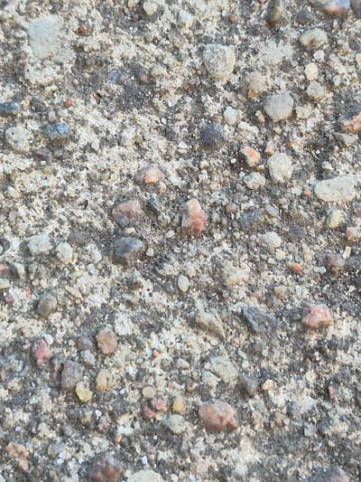 The image depicts a close-up, ground-level view of a patch of gravel and sand, likely found on a street or pathway in Švenčionys, Lithuania. The surface is composed of numerous small stones, pebbles, and coarse sand grains. The colors range from light gray and beige to darker shades of gray and brown, with occasional specks of reddish-orange from the stones. The texture appears rough and uneven, characteristic of an unpaved or weathered surface. No people, animals, or discernible structures are visible in the frame. The lighting suggests an outdoor environment, possibly under natural daylight. The scene is devoid of any specific landmarks or identifying features that would pinpoint a precise location within the city.
