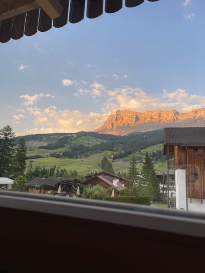The image displays a mountainous landscape with a village nestled in a valley. The sun is setting, casting a warm, golden glow on the rocky peaks of the mountains. The sky is a gradient of blue and orange, with scattered white clouds.
In the foreground, there are buildings with wooden architecture, suggesting a rustic and traditional setting. These buildings appear to be part of a village or resort, with some featuring balconies and outdoor seating areas. Lush green trees and rolling hills form the midground, leading the eye towards the majestic mountains in the background.
The scene evokes a sense of tranquility and natural beauty, characteristic of a mountain resort town during the golden hour. The clear sky and gentle light suggest pleasant weather.
There is no visible text in the image. The location context places this scene in La Ila - Stern - La Villa, Italy, a region known for its stunning alpine scenery.