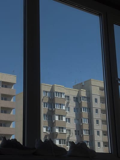 The image shows a view from a window. Outside the window, there are two buildings, one taller and one shorter, constructed from light-colored materials, likely concrete. Both have a repetitive pattern of windows and possibly air conditioning units. The sky is clear and blue. The photograph seems to be taken indoors, indicated by the window frame and the presence of a curtain on the right. The scene is daytime, and the architectural style suggests a post-Soviet construction era. Location is Shendzhii, Russia.