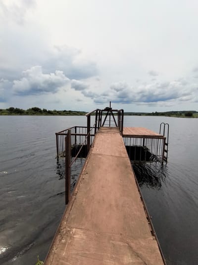 The image shows a wooden pier extending into a dark, choppy body of water. The pier is made of weathered wooden planks with metal railings. The background features a treeline with distant hills and a cloudy sky with shafts of light. The scene appears to be in a rural or natural setting, likely a lake or river. Based on the prompt, the location is specified as Кояново, Russia. There are no people or animals visible in the image. The overall atmosphere is overcast and slightly moody.