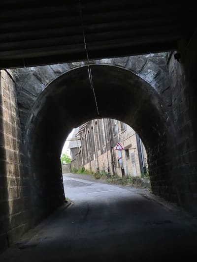 The image shows the view from within a dark, arched tunnel constructed of stone and brick. The tunnel curves, obscuring much of the view and creating a sense of enclosure. Outside the tunnel's opening, a street scene is visible. The perspective suggests the photograph was taken looking outwards from the tunnel.
The area outside the tunnel appears to be urban, with buildings constructed of light-colored brick visible in the distance. There is a road or pathway leading away from the tunnel, with some vegetation growing along the edges. A triangular road sign with a red border and a black arrow pointing to the right is mounted on a pole. The sky is overcast, suggesting a cloudy or possibly rainy day.
The lighting is dim within the tunnel, with some light filtering in from the outside. The overall mood is somewhat mysterious or dramatic due to the contrast between the dark tunnel and the brighter exterior. No people are visible in the frame. The setting appears to be in Käerjeng, Luxembourg, as indicated by the location context. The time of day is not definitively identifiable, but the overcast sky suggests it could be daytime. There are no other discernible text elements beyond the road sign.