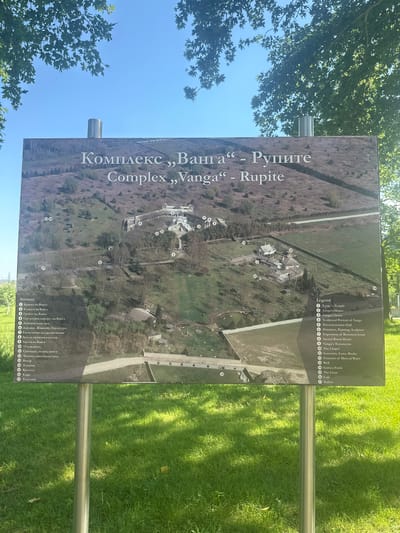 An outdoor informational sign, oriented vertically and featuring a map of the "Complex ,,Vanga'' - Rupite," is prominently displayed in what is identified as the area of General Todorov, Bulgaria. The sign is supported by two silver-colored, cylindrical metal poles embedded in vibrant green grass in the foreground, which exhibits patches of sunlight and shadow. Above the sign, a clear, bright blue sky is visible, partially framed by the lush green foliage of deciduous trees with distinct lobed leaves.
The sign itself is rectangular and presents an aerial perspective of the "Complex ,,Vanga'' - Rupite" on a textured, earthy-toned background depicting various buildings, pathways, and natural elements of the complex. The title is written in both Cyrillic script ("Комплекс ,,Ванга'' - Рупите") and Latin script ("Complex ,,Vanga'' - Rupite") at the top. A detailed legend is provided on the left and right sides of the lower portion of the sign, listing 17 numbered points of interest in both Bulgarian and English. These points include:
1. Храмът на Ванга (Vanga's Temple)
2. Къщата на Ванга (Vanga's House)
3. Гробът на Ванга (Vanga's Grave)
4. Скулптурен портрет на Ванга (Sculptural Portrait of Vanga)
5. Документална зала (Documentation Hall)
6. Дарения, Живопис, Скулптура (Donation, Painting, Sculpture)
7. Експозиция на дарени икони (Exposition of Bestowed Icons)
8. Дом за свещени ритуали (Sacred Ritual Home)
9. Завети на Ванга (Vanga's Testaments)
10. Параклисът (The Chapel)
11. Сувенири, икони, книги (Souvenirs, Icons, Books)
12. Чешма с минерална вода (Fountain of Mineral Water)
13. Бунар (Well)
14. Токачки (Guinea Fowls)
15. Кръстът (The Cross)
16. Кафe (Café)
17. Тоалетни (Toilets)
The map visually corresponds to these numbered locations, showing the layout of the complex with structures such as the temple and Vanga's house centrally positioned. The terrain depicted on the map includes undulating hills and sparse vegetation.