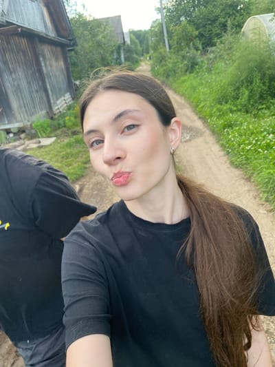 The media file is a portrait-style selfie taken outdoors, likely during the daytime as indicated by the natural lighting. The subject is a young person with long, brown hair, wearing a black t-shirt. Their face is turned towards the camera, with their lips pursed in a playful or pouting expression. Their eyes are open and looking directly at the lens.
The background suggests a rural or semi-rural setting. A dirt path is visible, flanked by lush green grass and vegetation. In the lower left portion of the image, a weathered wooden structure, possibly a shed or part of a house, is present. The overall scene evokes a casual, outdoor environment, potentially in a village or countryside.
No visible text is present in the media file. The time of day appears to be daytime, with soft lighting suggesting it might be late afternoon or early morning. The weather seems pleasant and conducive to outdoor activity. The expression of the subject implies a lighthearted or playful mood. The location context of Novyi Ural, Russia, further supports the interpretation of a rural or natural setting.