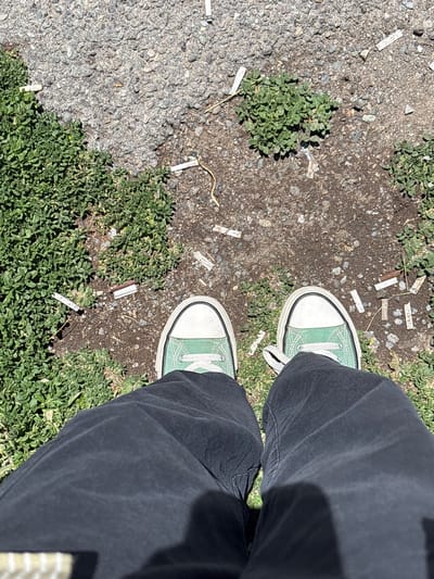 A first-person, high-angle shot shows a person's legs and feet standing on a patch of ground with gravel, dirt, and low-lying green vegetation. The person is wearing black pants and light green Converse sneakers. The ground is littered with numerous cigarette butts. The scene appears to be outdoors in a public area, possibly a sidewalk or roadside in the city of Jrarat, Armenia. The lighting suggests it is daytime.