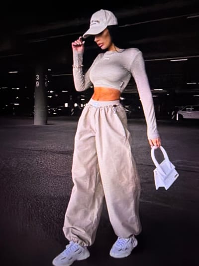 A woman stands in what appears to be a dimly lit, covered parking garage. She is wearing a light-colored crop top, matching baggy pants, a baseball cap, and athletic shoes. She is holding a small white handbag. The background shows concrete pillars, parked vehicles, and overhead lighting. There is no visible indication that this scene is located in Tomai, Moldova.