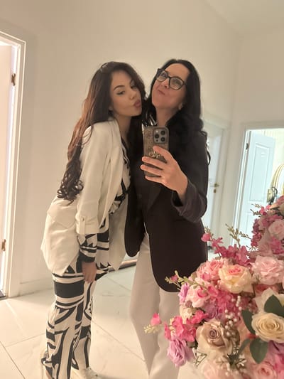 Two women pose for a selfie in front of a mirror with a bouquet of pink and white flowers. The older woman wears glasses and a dark blazer, holding a phone. The younger woman wears a white and black patterned outfit. They appear to be indoors, possibly in a hallway, based on the door frames visible in the mirror's reflection. The location is unknown and can not be inferred from the image.