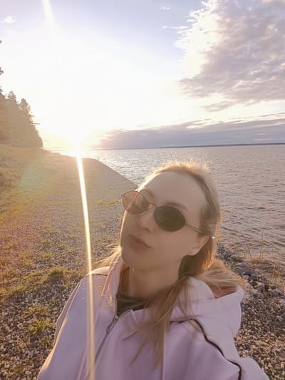 A woman takes a selfie outdoors in Вичелово, Russia, at sunset. She wears dark sunglasses and a light pink hooded sweatshirt. The background shows a shoreline with a body of water, possibly a lake, with visible waves. The setting sun creates a strong glare and is positioned near a distant shoreline. The ground near the woman is a mix of grass and gravel. Clouds are visible in the sky.
