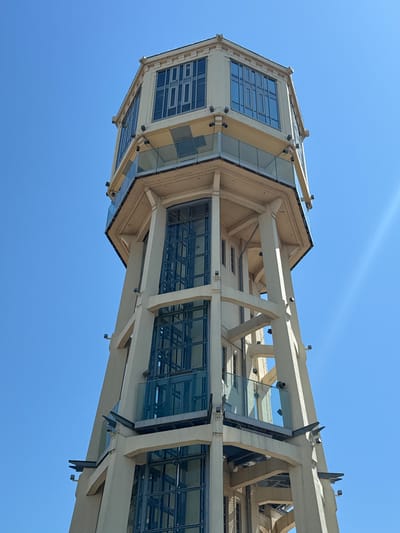 The image depicts the Siófok Water Tower, a landmark structure in the city of Siófok, Hungary, shot from a low angle looking upwards. The tower is constructed with a predominantly beige or light-colored framework made of support beams. The structure consists of multiple levels encased in glass. The sky appears as a clear, unblemished blue background. The architecture exhibits a modern design with geometric shapes and angles, featuring both solid construction and transparent glass sections.