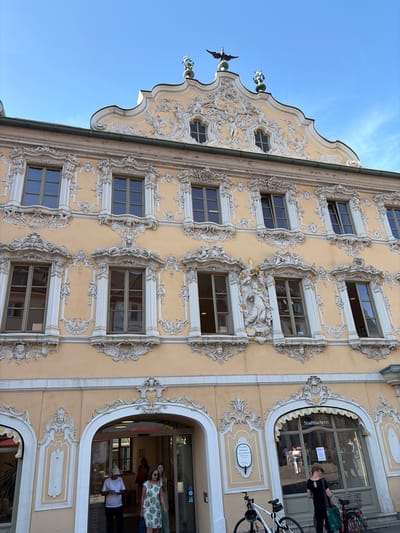 This image depicts the exterior of a historic building, identified as the Haus zum Falken (House to the Falcon), located at Marktplatz 2 in Würzburg, Germany. The building features an ornate Baroque facade painted in a pale yellow hue, adorned with extensive white stucco decorations. These decorations include intricate scrollwork, cherubs, floral motifs, and sculpted figures above and around the windows. The facade has multiple floors of windows, each framed by decorative elements.

In the foreground, an arched entrance leads into the building, with several people visible. To the right of the entrance, a storefront with the sign "Stadtbücherei" (City Library) is visible, with large windows reflecting the surroundings and people inside. Two bicycles are parked in front of the storefront.

The sky above is a clear blue with some wispy clouds. The architectural style and the specific building name place the scene in Würzburg.