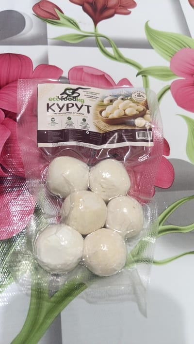 The image shows a vacuum-sealed package of kurut, a dried dairy product, which is a traditional Kyrgyz snack. The package is positioned on a surface with a floral print. The kurut balls are a light off-white to beige color and are individually wrapped in clear plastic within the larger sealed package. The packaging has text and images, including a picture of kurut in a bowl and traditional yurts, suggesting a connection to Kyrgyz culture. The brand name "ecofoodkg" is visible. The surface it rests on has large, stylized pink and green floral designs. The specific location in Balykchy, Kyrgyzstan cannot be determined from the image alone.