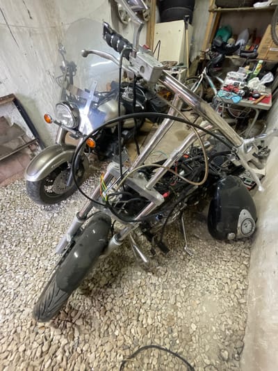 The image captures a partially disassembled or stored motorcycle within an indoor garage or workshop setting in Znamensk, Russia. The motorcycle, primarily chrome and black, is oriented diagonally across the frame, resting on a gravel floor.
The main subject is a cruiser-style motorcycle. Prominent features include wide chrome handlebars, a black fuel tank with a chrome-ringed speedometer/gauge cluster, and a large, clear windshield. A single round headlight and orange front turn signals are visible. The front wheel features a disc brake, and its tire shows a distinct tread pattern. A grey or silver front fender partially covers the wheel. Exposed wires, some with colorful insulation, are visible near the handlebars and steering column, indicating the motorcycle is either in a state of repair, customization, or long-term storage.
The background consists of shelves cluttered with various items, including cardboard boxes, tools, plastic containers, and what appears to be a bicycle frame. This suggests a functional, possibly disorganized, storage or work environment. The floor throughout the scene is covered in small, light-colored gravel.
No people are present, and no active event or work is taking place. The motorcycle is stationary, implying it's a project, awaiting parts, or simply being stored. The lighting is artificial, leaving the time of day indeterminate. On the brake fluid reservoir cap on the left handlebar, the text "DOT" is visible.