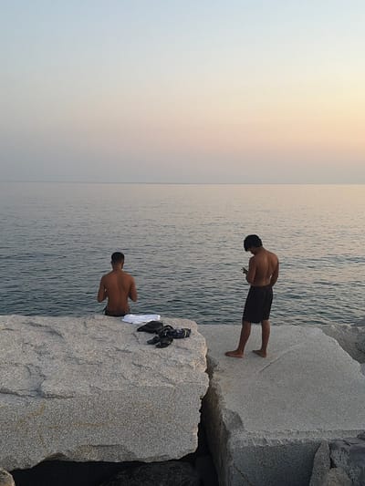 The image shows two young men standing on large granite rocks next to the sea. The scene is set during what appears to be late afternoon or early evening, as indicated by the soft, warm glow in the sky, transitioning from a pale yellow to a soft blue. The water is calm with gentle ripples. The man in the foreground is facing away from the camera, looking down at the water. He is wearing dark shorts. Behind him and slightly to the left, another man is also facing away from the camera, his head bent. He is wearing dark shorts as well. On the rock next to the man in the foreground, a white towel and a black bag are placed. The setting is a rocky coastline, possibly a breakwater or a natural formation of large boulders by the sea. The atmosphere appears serene and peaceful, with no text visible in the image. The location is identified as Imperia, Italy.