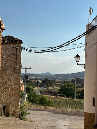 The image depicts a street view in the village of La Cuevarruz, Spain. On the left, a rough-hewn stone building with a red-tiled roof and a barred window occupies the foreground. Overhead, several dark electrical wires stretch across the frame. To the right, a cream-colored building with a decorative black wrought-iron lamppost attached to its exterior is visible. The street slopes downwards and curves to the right, leading towards a rural landscape. This landscape features scattered houses, trees, fields, and a winding road in the middle ground, with rolling hills and mountains visible in the distance under a clear blue sky with some scattered clouds. A tall electrical pole with a transformer is situated in the middle ground, and a black metal fence runs along the edge of the property on the right. The scene appears to be illuminated by daylight.