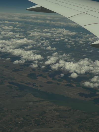 This is an aerial photograph taken from an airplane window, looking down at clouds and the landscape below. The airplane wing is visible in the upper right corner of the frame. The clouds appear fluffy and white, casting shadows on the land. The terrain below is a mix of arid land and agricultural fields, with some circular patterns that suggest irrigation systems. A body of water, possibly a river or lake, runs through the landscape. The scene is illuminated by sunlight. The image does not provide any specific information about the city of Panos, Turkey.