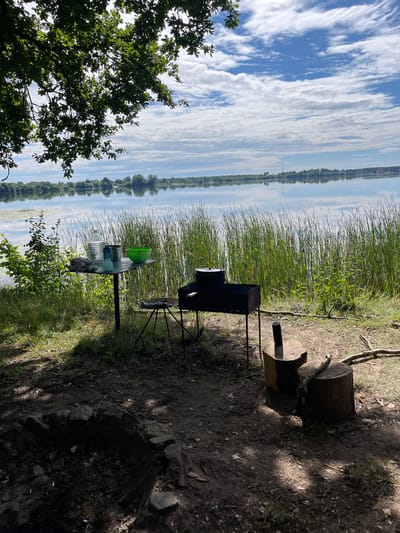 The image depicts a lakeside campsite in the Вяззеўскі сельскі Савет, Belarus. In the foreground, a makeshift fire pit is visible, surrounded by a dirt and grass area. To the right, two tree stumps serve as seating, with a branch resting on one. A portable grill with a pot on top stands near the stumps. Further to the left, a small table holds several items, including a large bottle of water, two smaller bottles, a metal container, and a green bowl. Behind the table, tall reeds line the edge of a calm lake. The lake reflects a bright blue sky filled with scattered white clouds. The background shows a treeline and distant buildings on the opposite shore. Lush green foliage, including tree branches with leaves, partially frames the top of the image. The overall scene suggests a peaceful outdoor setting, possibly for a picnic or camping trip.