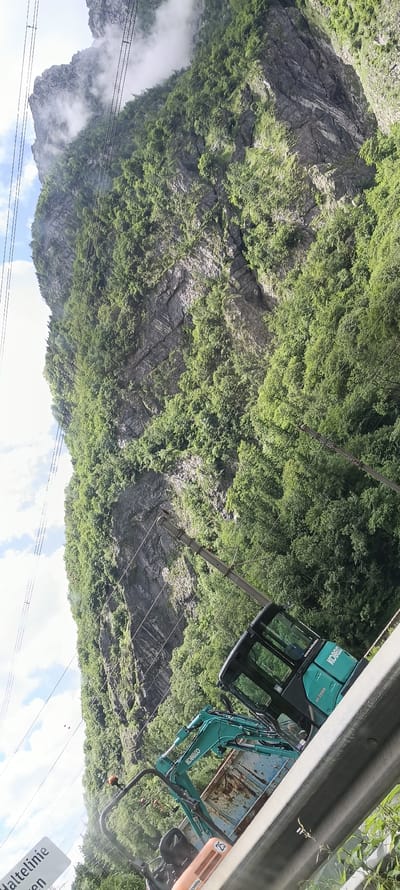 In Golling an der Salzach, Austria, a rugged mountain slope covered in lush green vegetation dominates the scene. A sheer rock face, streaked with gray and white, is visible beneath the foliage. Overhead, a network of power lines stretches across the frame, supported by metal poles. In the foreground, a turquoise excavator is parked, its cab featuring the "KOBELCO" logo. A rusted truck bed is attached to the excavator. To the left, a white sign with German text, including "Haltelinie" and "en," is partially visible. The sky above is a mix of white clouds and blue patches. The image is oriented vertically, with the mountain occupying the majority of the frame.