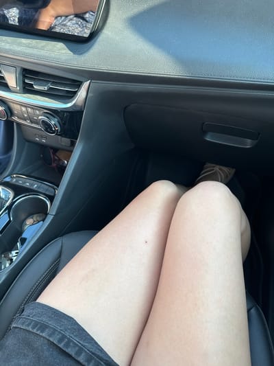 The image shows a person's legs from the thigh down, seated in the passenger seat of a car. The person is wearing denim shorts and sandals. The interior of the car is visible, including the dashboard, air vents, and a cup holder containing coins. A pair of sunglasses rests on the floor of the passenger side. The scene appears to be a daytime shot, possibly on a sunny day, given the lighting. The focus is on the legs and the immediate car interior, with no specific activity or event clearly depicted. No visible text is present in the image.