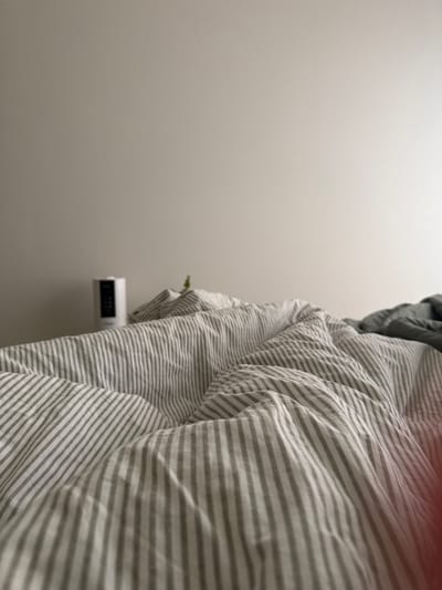 The image shows a bedroom with a striped duvet cover dominating the right side of the frame. A white digital clock is mounted on the gray wall to the left of the bed. The rest of the left side of the image is a plain gray wall. The location, Люк, Russia, is not discernible from the visual elements.
