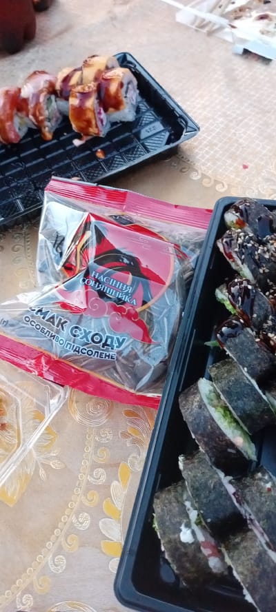 The image shows a table setting with several portions of sushi and a bag of snacks, likely in Krasivka, Ukraine. The table is covered with a patterned tablecloth. In the foreground, on the left, is a clear plastic container with a small amount of food, possibly a dipping sauce or condiment. To the right of this container, and partially obscuring it, is a red and white plastic bag with Cyrillic text that reads "НАСІННЯ СОНЯШНИКА" (Sunflower Seeds) and "МАК СХОДУ ОСОБЛИВО ПІДСОЛЕНЕ" (Poppy Seeds Especially Salted).

The main subjects are two black plastic trays of sushi. The tray on the left contains several pieces of sushi with a glossy, reddish-brown topping and white sauce. The tray on the right contains sushi rolls wrapped in seaweed, sprinkled with sesame seeds and drizzled with a dark sauce. The sushi appears to be prepared and ready for consumption.

The environment suggests an outdoor or casual indoor setting, given the presence of disposable containers and the general presentation. The lighting indicates it's daytime. No people are visible in the image. The overall scene depicts food ready to be eaten.