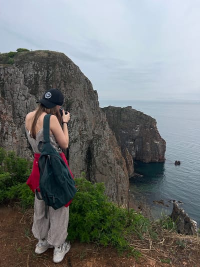 A person stands on a cliff's edge, presumably in Russky Island, Russia. The person is looking through a camera. They are wearing a black baseball cap, a checkered dress, a green backpack, and white sneakers. A red jacket is visible beneath the backpack. The rocky cliff falls down to the ocean, with an additional large rock formation in the water. The sky is overcast. Vegetation grows along the cliff's edge.
