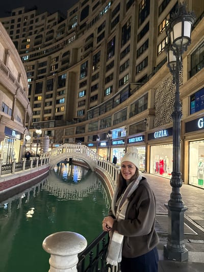 This nighttime image depicts an urban environment in Esenler, Turkey, specifically a themed shopping and residential complex designed with Venetian architectural influences. The scene is dominated by a central female subject and features a canal, a bridge, and multi-story buildings.

The foreground is occupied by a female individual, approximately 20-30 years old, positioned slightly to the right of the frame's center. She is smiling and looking directly at the camera. She wears a white knit beanie, a grey and white plaid scarf, a dark brown hooded sweatshirt, and dark blue pants. Her hands are clasped over a black metal railing with a white stone post, which separates her from a narrow, artificial canal.

The canal, filled with still, greenish-blue water, occupies the midground. The water reflects the overhead artificial lights and the illuminated shopfronts. A prominent white, stone-arched bridge with balustrades spans the canal in the middle-left of the image. On the right side, an ornate black street lamp with two globes stands vertically, partially obscuring a shopfront.

The background consists of tall, curved, multi-story buildings that line both sides of the canal. These structures feature numerous windows, many of which are illuminated, indicating occupancy. The ground floor of these buildings hosts various retail stores with brightly lit window displays and visible signage, including "GIZIA," "TAMER TANCA," "celco," and "UNESCOL." Mannequins displaying clothing are visible in several shop windows. Decorative architectural elements, such as textured patterned walls and classical columns, are also present. Several other indistinct figures are visible on walkways and near shop entrances in the background. The sky above is dark, indicating a nighttime setting.