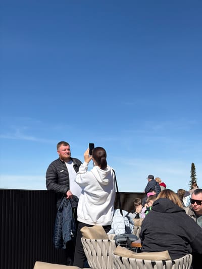 The image shows a group of people on an elevated platform. A man in a black jacket is being photographed by a woman in a white hoodie. Other people, including children, are seated on beige chairs in the background. A tall evergreen tree is also visible. The sky is clear blue. The location is in or near Ozernoe, Russia.