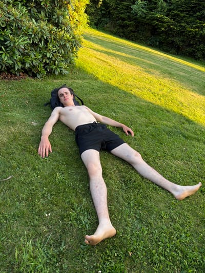 A young man with dark hair, wearing black shorts, lies on his back on a grassy lawn. His arms are outstretched to his sides, and his legs are spread apart. He appears to be looking up at the sky. Behind him, a large green bush and a line of dark green trees form a backdrop. Sunlight casts long shadows across the lawn, suggesting it is late afternoon or early evening. The scene is peaceful and appears to be set in a park or a garden. The location is identified as Mordarka, Poland.